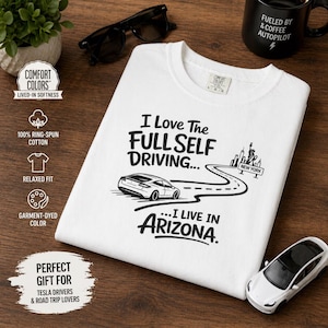 以下が含まれることがあります： 「I Love The Full Self Driving... I Live In Arizona」のテキストが入った白いTシャツ。ニューヨークのスカイラインと道路上の車のグラフィックが特徴です。シャツにはComfort Colorsのラベルも付いています。