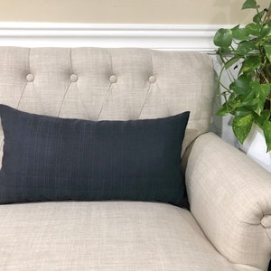 May include: A black rectangular pillow with a textured surface sits on a light beige tufted sofa.