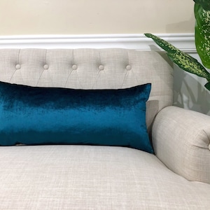 May include: A teal blue velvet lumbar pillow on a light beige couch.