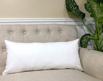Könnte beinhalten: Ein weißes rechteckiges Kissen auf einem beigefarbenen, gesteppten Sofa mit einer grünen, belaubten Pflanze im Hintergrund.