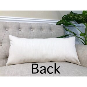 May include: A plain white rectangular cushion with a subtle texture, placed on a light grey tufted sofa. The word "Back" is printed in black below the cushion.