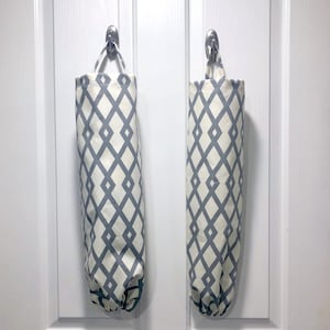 May include: Two fabric bag dispensers hanging on a white door. The bags are cylindrical with a cream background and a repeating gray diamond pattern. Each bag has a loop at the top for hanging.