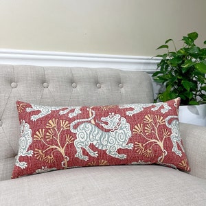May include: A red lumbar cushion with a white and blue stylised lion design. The cushion has a red background with a subtle texture and a white and blue lion design with a yellow tree branch pattern.