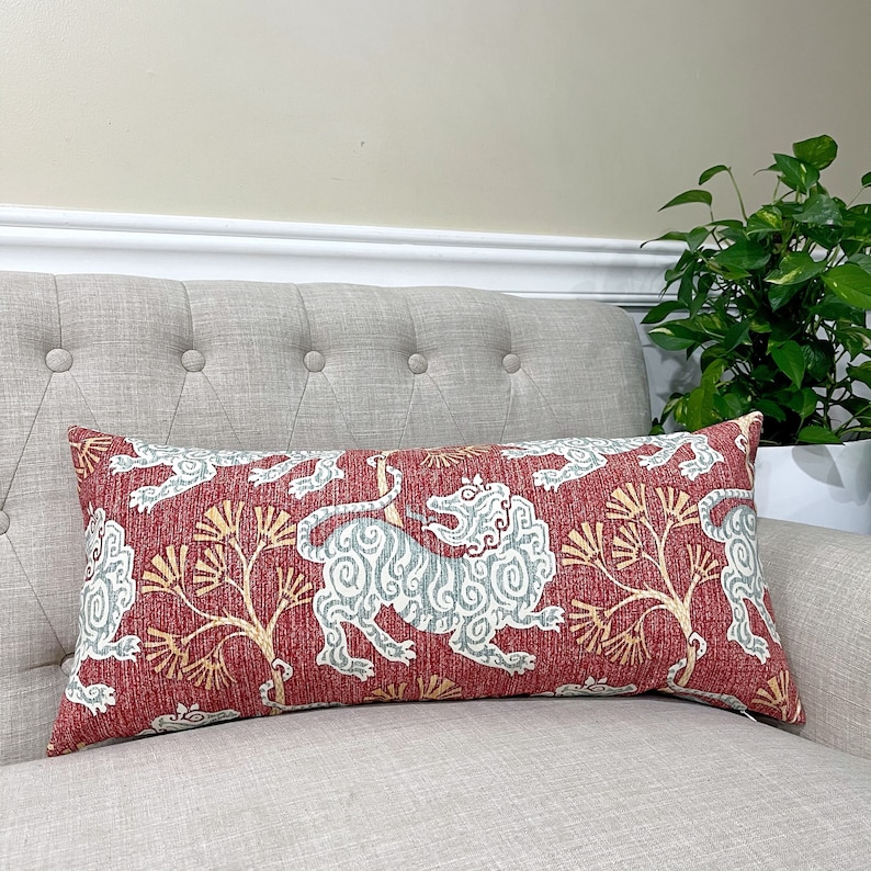 May include: A red lumbar cushion with a white and blue stylised lion design. The cushion has a red background with a subtle texture and a white and blue lion design with a yellow tree branch pattern.