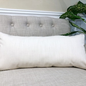 May include: A rectangular, off-white lumbar pillow with a textured linen-like appearance. The pillow is placed on a light gray upholstered chair with button tufting. A green plant is visible in the background.