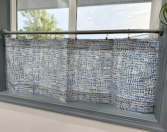 May include: A blue and white patterned valance curtain hanging in a window. The curtain has a repeating dot design. The curtain is hung on a metal rod with metal rings. The window frame is gray.