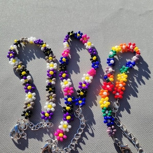 May include: Three beaded bracelets with flower-shaped beads. The bracelets are in the colors of the non-binary, pride, and genderfluid flags.