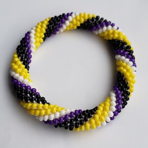 May include: A beaded bracelet in a non-binary pride flag design. The bracelet features alternating rows of yellow, white, purple, and black beads. The bracelet is circular and appears to be handmade.