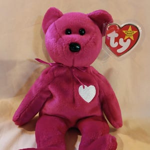 May include: A bright magenta plush teddy bear with black plastic eyes and a black nose. The bear features a small white fabric heart stitched onto its chest and a matching magenta ribbon tied around its neck. A heart-shaped Ty Beanie Baby tag is attached.