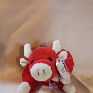 May include: A plush toy bull with bright red fur and white accents on the horns, ears, snout, and hooves. The bull has small black eyes and a clear plastic tag attached to its side, featuring printed text and a barcode on the visible surface.