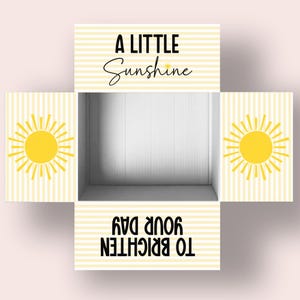 Puede incluir: Una caja blanca con rayas amarillas y blancas. La parte superior de la caja dice "A Little Sunshine" y la parte inferior dice "To brighten your day". Hay un sol amarillo a cada lado de la caja.