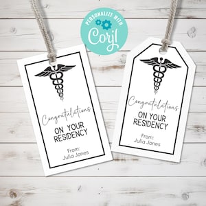 以下が含まれることがあります： 黒い縁取りと黒いカドゥケウスのシンボルが付いた白いギフトタグが2つ。タグには「レジデンシーおめでとう」と「From: Julia Jones」の文字が書かれています。