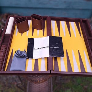 Puede incluir: Un juego de backgammon de cuero marrón con un tablero de juego amarillo y blanco, un juego de fichas y un libro de reglas. El juego está abierto y listo para jugar.