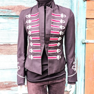 May include: Black military-style jacket with red and white trim. The jacket features a high collar and decorative white accents on the sleeves. The front has vertical red stripes with white detailing. The jacket is worn over a black turtleneck.