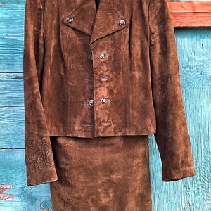 A brown suede jacket and skirt set with embroidered details. The jacket has a mandarin collar and a double breasted closure. The skirt is a-line and hits above the knee. It is shown hanging on a hanger