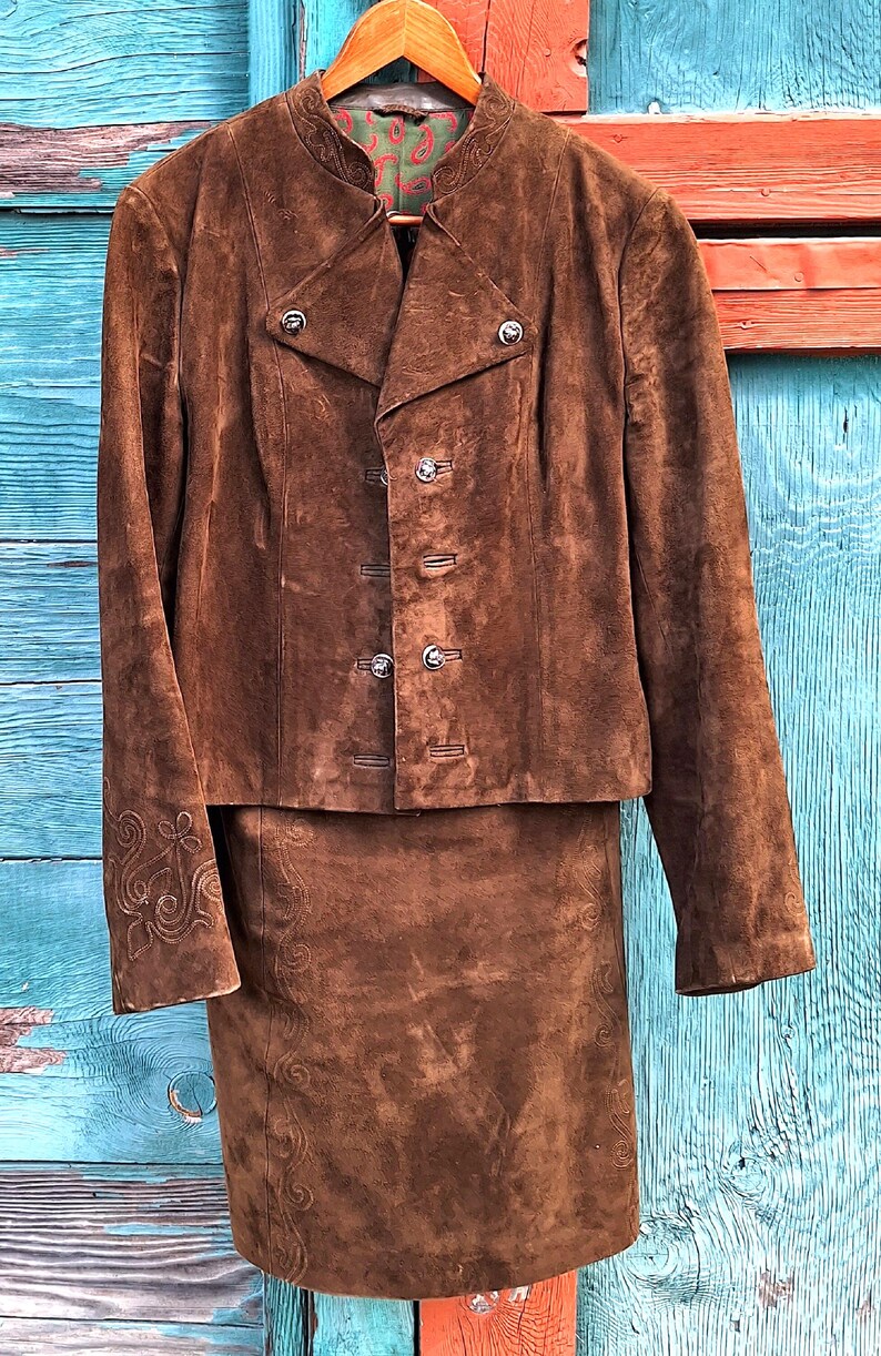 A brown suede jacket and skirt set with embroidered details. The jacket has a mandarin collar and a double breasted closure. The skirt is a-line and hits above the knee. It is shown hanging on a hanger