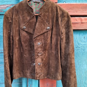 A brown suede jacket with a mandarin collar and silver buttons. The jacket is hanging on a wooden hanger against a blue and orange wooden background.