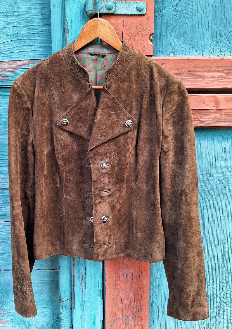 A brown suede jacket with a mandarin collar and silver buttons. The jacket is hanging on a wooden hanger against a blue and orange wooden background.