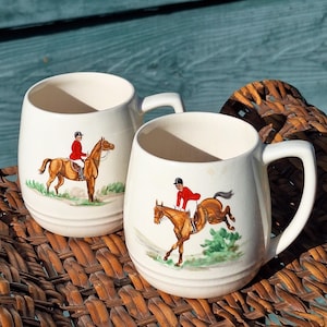 Puede incluir: Dos tazas de cerámica blanca con un diseño de caballo y jinete marrón. Las tazas tienen una forma redondeada y un asa en el lado derecho. El jinete lleva una chaqueta roja y un sombrero negro. Una taza presenta un caballo saltando una valla, mientras que la otra taza presenta un caballo caminando.