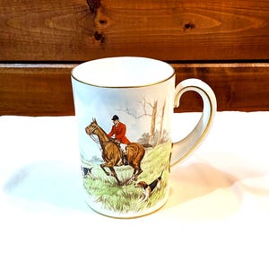 Puede incluir: Taza de cerámica blanca con borde y asa dorados. La taza presenta una escena de caza con un jinete a caballo marrón, perros y un paisaje. El asa es curva y tiene detalles dorados.