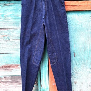 A pair of dark blue denim riding pants with knee patches. The jeans are hanging on a wooden hanger from a hook .