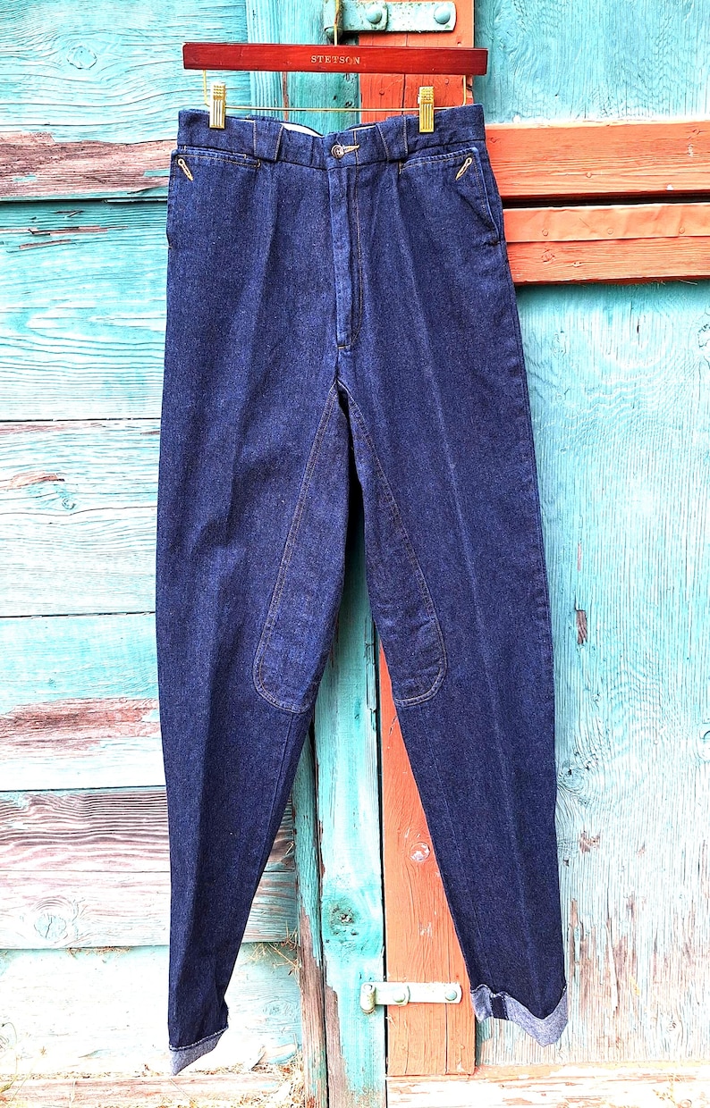 A pair of dark blue denim riding pants with knee patches. The jeans are hanging on a wooden hanger from a hook .