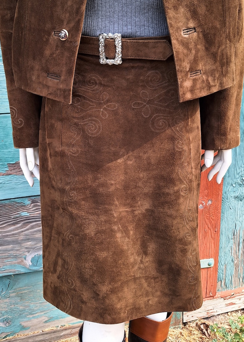 A close up view of a brown suede skirt with embroidered details. The skirt has a brown leather belt with a silver buckle.