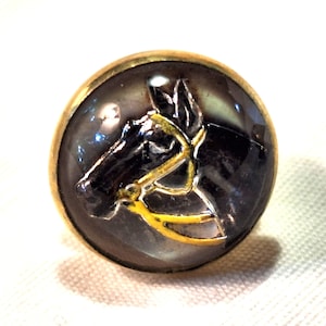 May include: A round, gold-toned button with a black horse head design. The horse is wearing a yellow bridle.