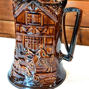 May include: A brown ceramic beer jug with a detailed relief design. The jug features a house-like structure and figures on horseback. The handle is a dark, glossy brown. The overall design is reminiscent of a rustic, vintage style.