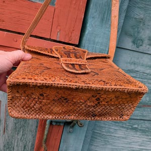 Bottom of a structured snakeskin handbag in the colors of chestnut and black. The front has a tab closure and it has a long shoulder strap The bottom is slightly flared with stitching detail on the edges.