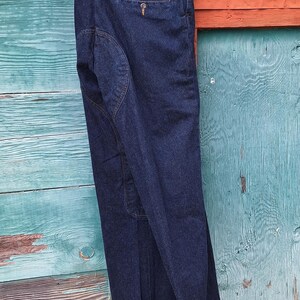 A pair of dark blue denim riding pants with a cuffed hem. The jodhpurs are hanging on a wooden hanger against a blue and orange wooden background.