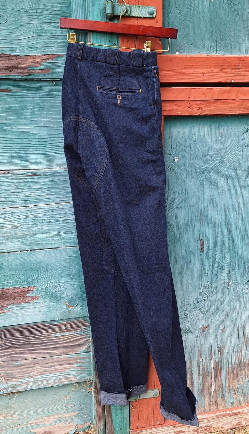 A pair of dark blue denim riding pants with a cuffed hem. The jodhpurs are hanging on a wooden hanger against a blue and orange wooden background.