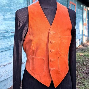Op de afbeelding: Een oranje suède vest met vier knopen en twee voorzakken. Het vest wordt getoond op een mannequin met een zwarte coltrui. Het vest heeft een levendige oranje kleur.