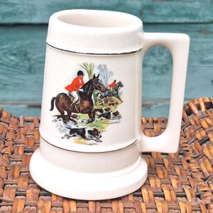 May include: Cream-colored ceramic beer stein with a handle and a detailed illustration of a fox hunt scene. The illustration features riders on horseback, hounds, and a landscape background. The stein has a gold trim.