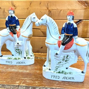Peut inclure: Deux figurines de chevaux en céramique blanche avec des cavaliers en vestes bleues et chapeaux rouges. Les figurines portent l'inscription "FRED ARCHER" sur la base. Les chevaux ont des accents dorés et sont tournés l'un vers l'autre.