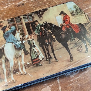 Puede incluir: Lata rectangular vintage con una escena pintada de personas a caballo con ropa de época. La obra de arte presenta a un hombre con abrigo rojo y a un hombre con abrigo azul. La lata tiene un borde azul y está sobre una superficie de madera.