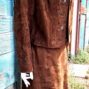 A brown suede jacket and skirt set with embroidered details. The jacket has a stand-up collar, a button closure, and two flap pockets. The skirt is a-line and has a straight hem.