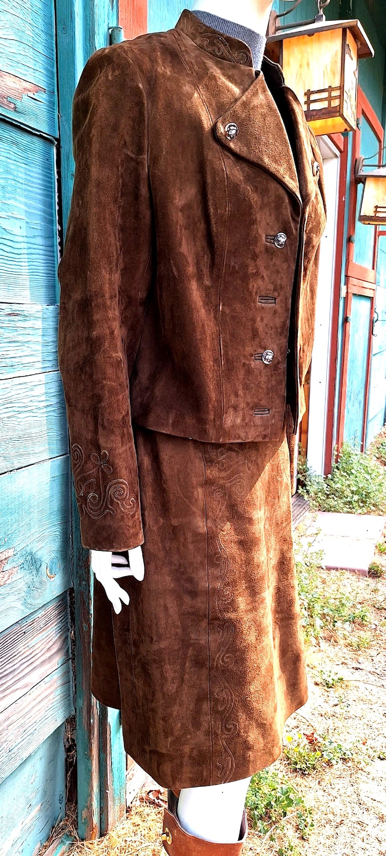 A brown suede jacket and skirt set with embroidered details. The jacket has a stand-up collar, a button closure, and two flap pockets. The skirt is a-line and has a straight hem.