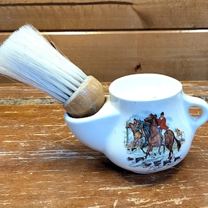 Puede incluir: Un cuenco de afeitado de cerámica blanca con una ilustración de una escena de caza y una brocha de afeitar con mango de madera. La brocha tiene cerdas blancas. El cuenco tiene un asa y un pico.