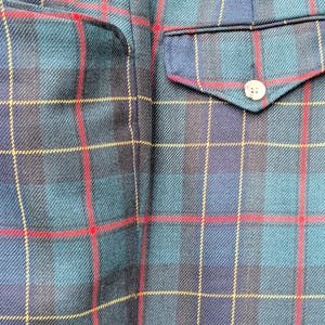 May include: Close-up of a section of a plaid fabric, featuring a pattern of intersecting lines in teal, navy, red, and yellow. The image shows a pocket with a button and a portion of a seam, suggesting a garment.
