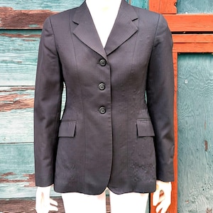 Puede incluir: Un blazer negro carbón con un corte entallado, con solapa de muesca, cierre de tres botones y dos bolsillos delanteros. La chaqueta tiene un sutil patrón de rayas verticales y se muestra en un maniquí.