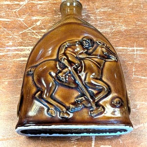 May include: Brown ceramic flask with a detailed relief of a person on a horse, likely depicting a polo player. The flask has a rounded shape with a white base and a cork stopper.