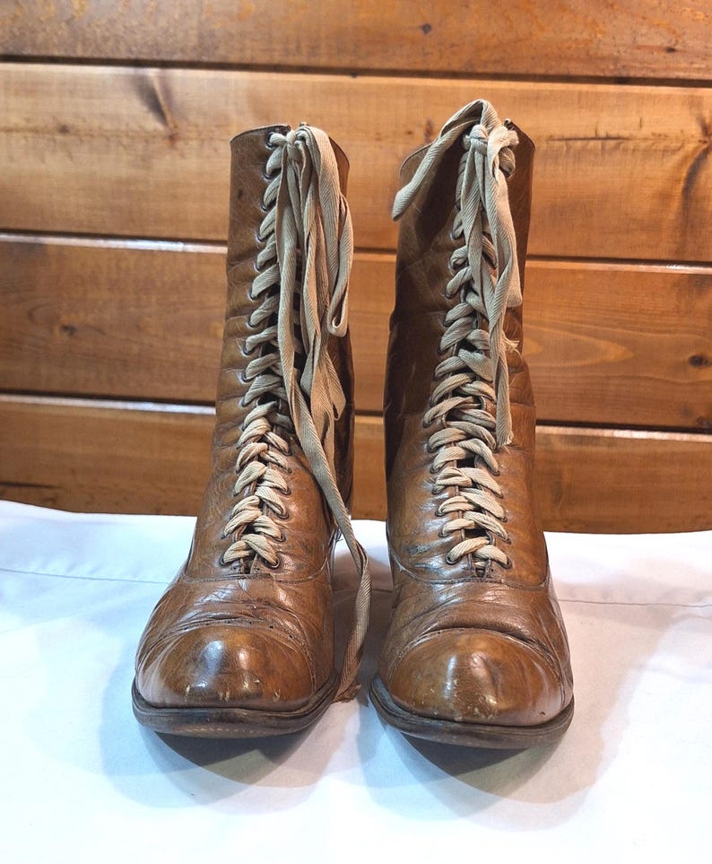 Front view of a pair of brown leather ladies Victorian boots with low heels and tan laces. The boots are narrow and long with above the ankle height