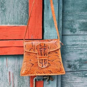 Może przedstawiać: Pomarańczowa torba na ramię ze wzorem skóry węża, zamykana na klapę i z długim, cienkim paskiem. Torba posiada ozdobną klamrę i wykonana jest z teksturowanego materiału. Torba wisi na tle zwietrzałego drewnianego tła.