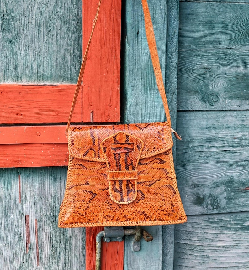 A brown snakeskin purse hanging from a hook against a green and orange barn side. The purse has a long strap and a tab front closure. The colors are chestnut and black python leather