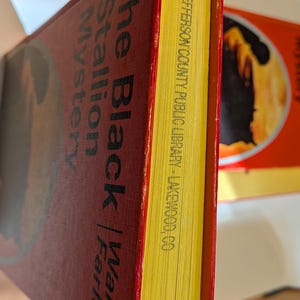 A hardcover book titled "The Black Stallion Mystery" by Walter Farley. The book has an orange/red cover with black lettering and yellow-trimmed pages. The pages are stamped with "JEFFERSON COUNTY PUBLIC LIBRARY - LAKEWOOD, CO."