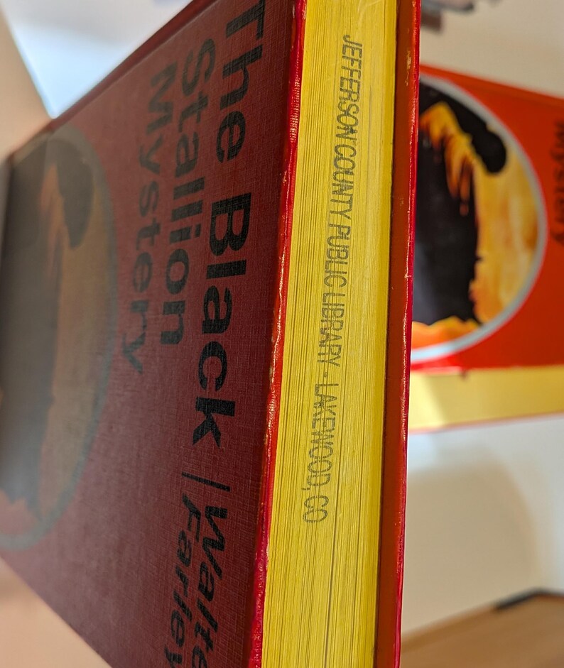 A hardcover book titled "The Black Stallion Mystery" by Walter Farley. The book has an orange/red cover with black lettering and yellow-trimmed pages. The pages are stamped with "JEFFERSON COUNTY PUBLIC LIBRARY - LAKEWOOD, CO."