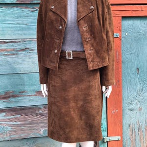 A brown suede skirt and jacket set with a decorative embroidered pattern. The skirt has a belt with a silver buckle. The jacket has a high collar and button closures.