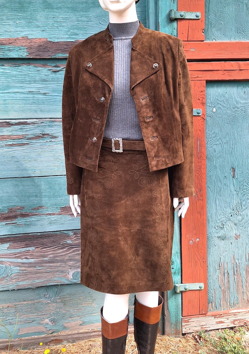 A brown suede skirt and jacket set with a decorative embroidered pattern. The skirt has a belt with a silver buckle. The jacket has a high collar and button closures.