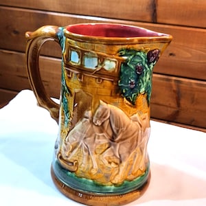 May include: A brown ceramic pitcher with a handle and a scene of horses pulling a carriage. The pitcher has green, brown, and white details.